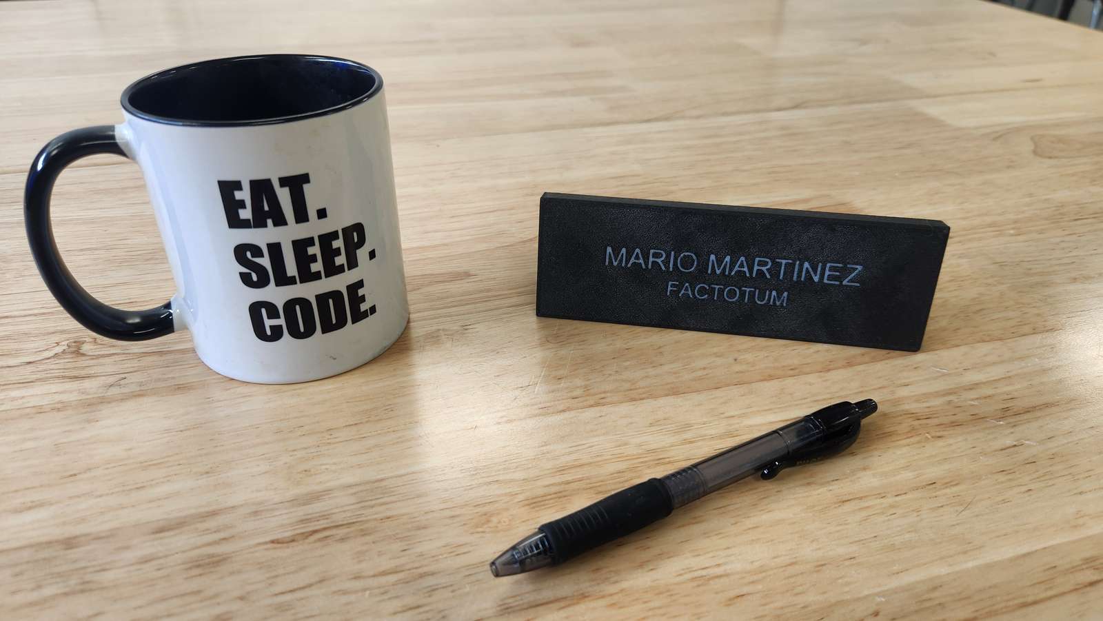 A black 3D-printed desk nameplate reading Mario Martinez, Factotum, on a wood desk beside a coding mug and a pen.